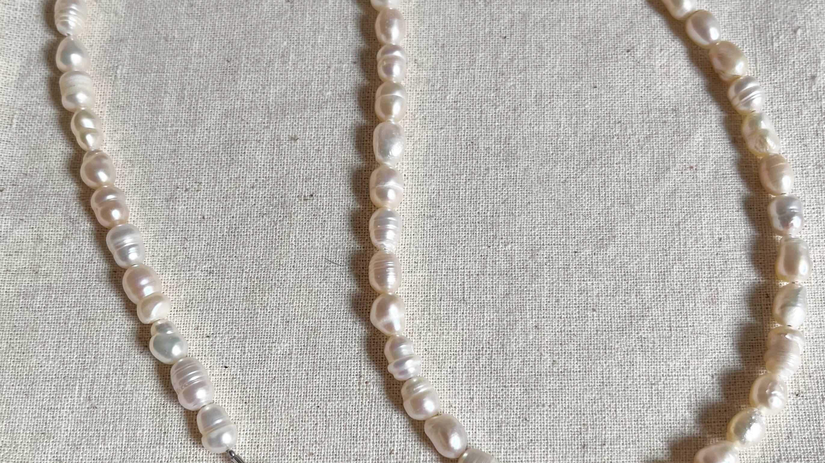 Cadenas de perlas blancas irregulares sobre tela beige, con broche plateado visible