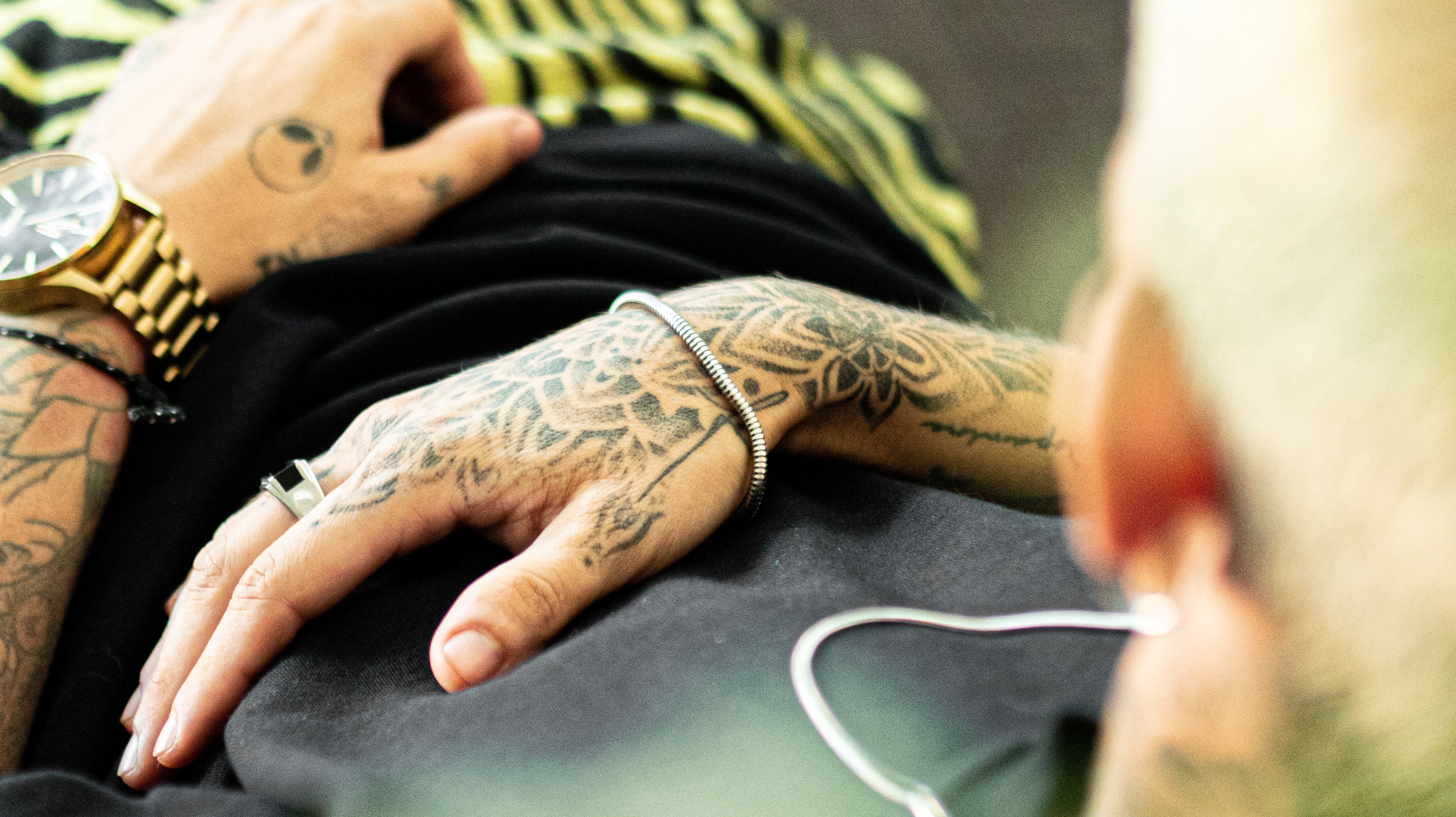 Tattooed arm resting on a surface showing a gold wristwatch and a silver bracelet.