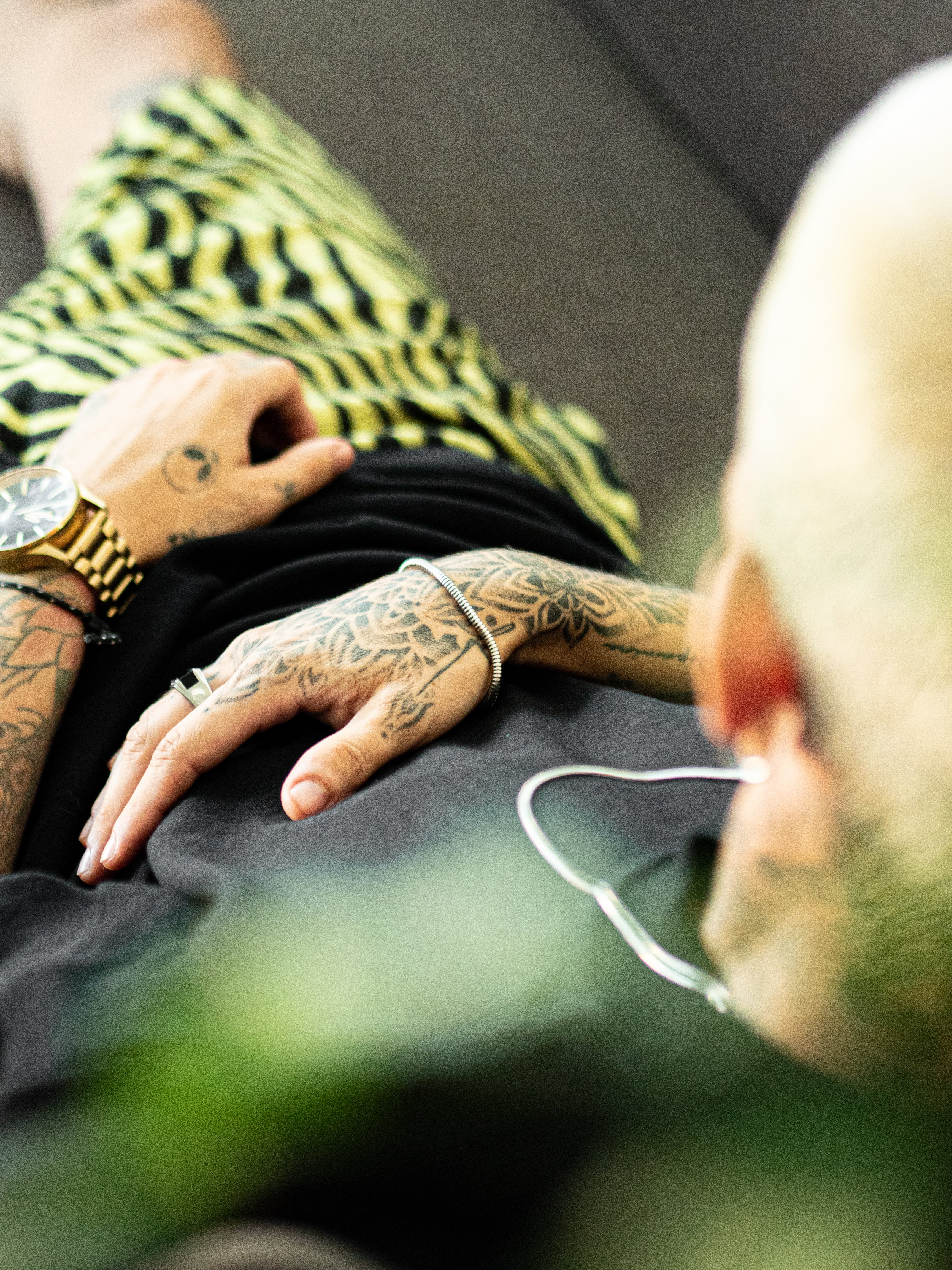 Tattooed arm resting on a surface showing a gold wristwatch and a silver bracelet.