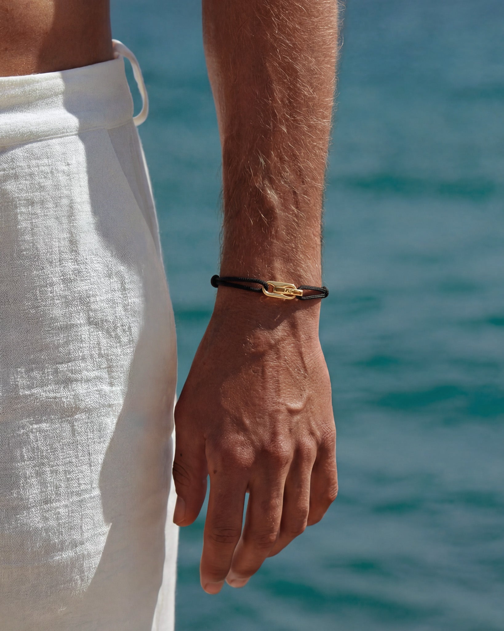 Wrist with a black cord bracelet and gold clasp against a seaside background.