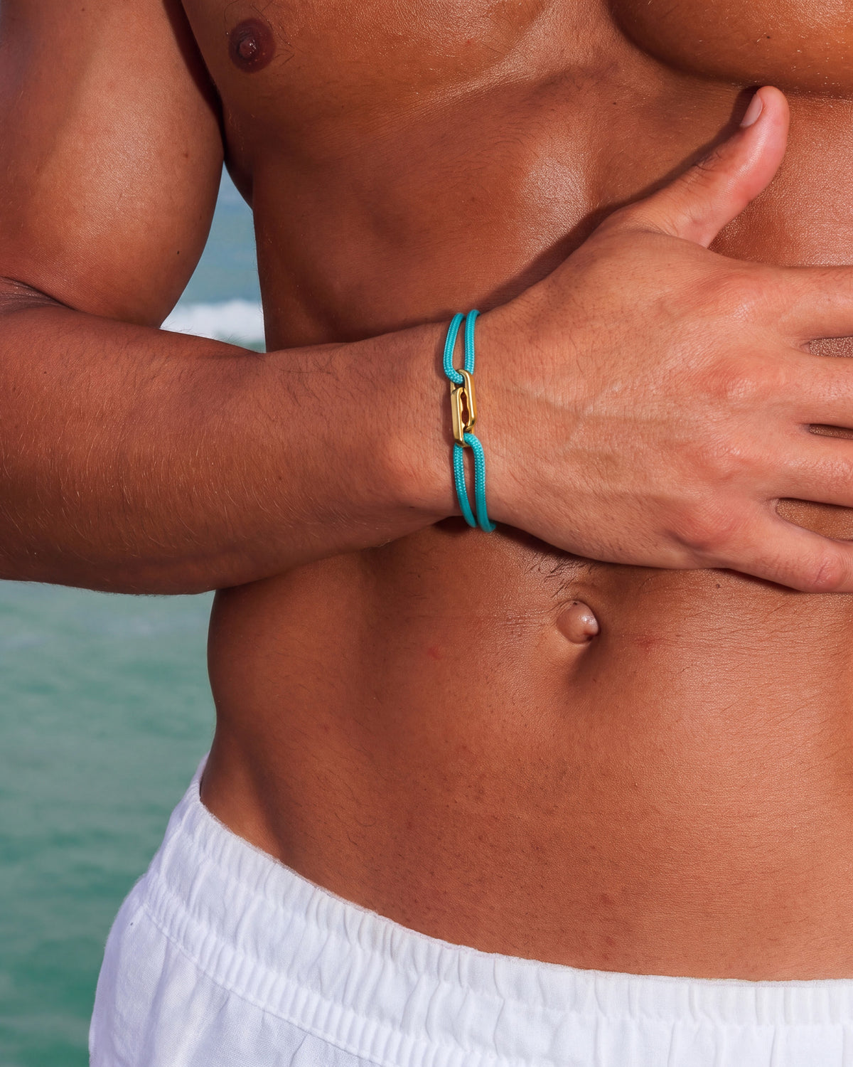 Atlantis gold bracelet with turquoise cord worn on the wrist of a shirtless man at the beach